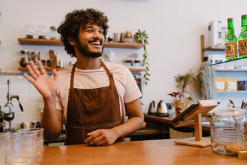 Fototapeta na wymiar Young indian smiling handsome curly barista standing behind the counter