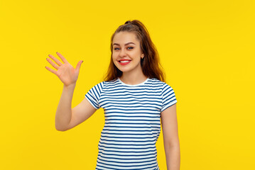 Friendly cheerful brunette girl smiling, saying hi and waving hand to greet person, make hello gesture, welcome someone, standing in white-blue striped t shirt over yellow background