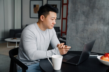 Asian man working with laptop while drinking coffee in kitchen