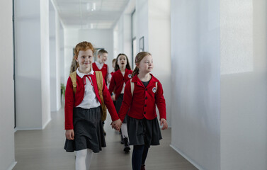 Happy schoolgrirl with Down syndrome in uniform holding hands with her classmate walking in scool...