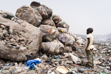 African child in a smoky landfill looking at giant bags with plastic bottles and household items to...