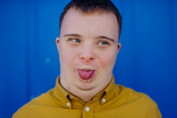 Close-up of happy young man with Down syndrome grimacing and sticking out tongue against blue...