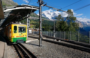 Beautiful scenery of a local train parking by the platform of Wengen Station with majestic snow capped Jungfrau mountain in background on a cloudy summer day in Bernese Oberland, Switzerland, Europe