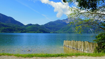 malerischer Blick über Kochelsee in Bayern mit Gras, Palisaden, Bergen, blauem Himmel und weißen...