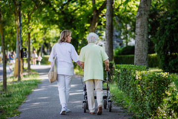 Rear view of caregiver with senior woman on walk in park with shopping bag.