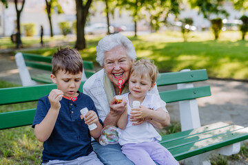 Great grandmother sitting on bench with her grandchildren and blowing soap bubbles together,...