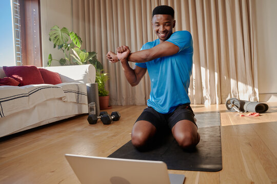 Young black African American happy working out at home stretching his arms before doing an online exercise class on his yoga mat in his modern apartment