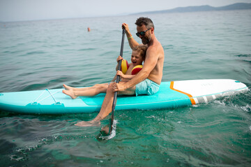 Happy father and little son on paddle board in sea together. Fatherhood and quality time with child concept.