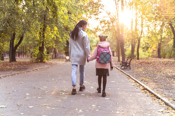 Mom and daughter walk in autumn park. Little girl after school with mother from back. Everyday life