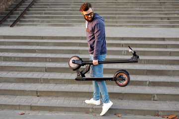 Man Carry Electric Scooter In Folded Position. Guy Carrying folded E-Scooter in hand on stairs. Male wear stylish clothes and hold electric scooter.