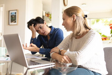 Caucasian mid adult lesbian woman looking at stressed girlfriend with head in hands sitting at table