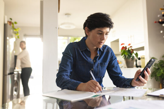 Caucasian mid adult lesbian woman with short hair using mobile phone and writing in book on table
