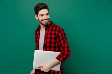 Young happy smiling cheerful fun cool man he 20s in red shirt grey t-shirt hold closed laptop pc computer look aside on workspace area mock up isolated on plain dark green background studio portrait