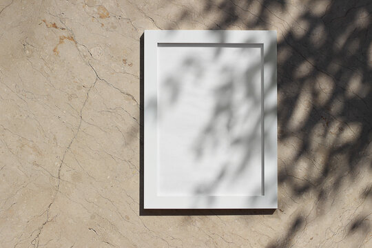 Blank white picture frame against beige marble background in sunlight. Dark tree leaves, branches silhouettes overlay, harsh shadows. Empty poster mockup for art display. Minimal summer design.