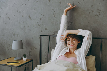 Young calm cheerful woman wear white shirt pajama she lying in bed rest relax spend time in bedroom...