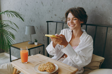 Young woman wear white shirt pajama she lying in bed use mobile cell phone take photo of breakfast on table rest relax spend time in bedroom lounge home in hotel room wake up dream in good mood day.