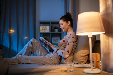 people, bedtime and rest concept - happy teenage girl reading book in bed at home at night