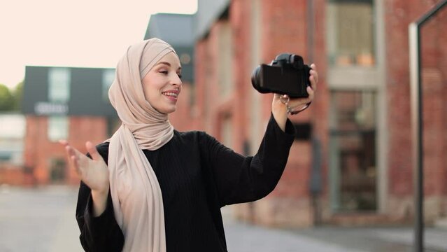 Young muslim woman using a video camera to record journey with smile. Happy and excited 20s arabic girl traveler wear black clothes with hijab, waving to internet audience. Influencer vlogging concept