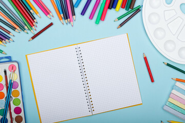 Blank sheet of paper of an open math notebook, art school supplies for painting, on a blue background with copy space for text. Colorful pencils, markers, paints, crayons. Top view. Back to school