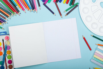 Blank sheet of paper of an open math notebook, art school supplies for painting, on a blue background with copy space for text. Colorful pencils, markers, paints, crayons. Top view. Back to school