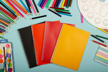 Multicolored notebooks, black, red and orange, art school supplies for painting, on a blue background with copy space for text. Colorful pencils, markers, paints, crayons. Top view. Back to school