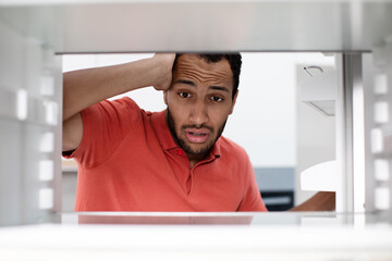 Starving hungry shocked disappointed young african american man checks empty fridge got stress in...