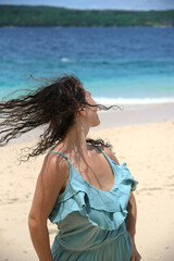 Fototapeta na wymiar A beautiful woman in a blue dress and with dark curly hair is having fun on the beach. Tourist vacation in a tropical resort. A cheerful girl with a curly hairstyle. Free lifestyle. Back view