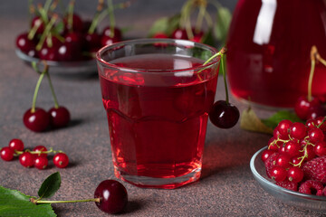 Homemade compote of red berries such as cherries, raspberries and currants in a glass and jug on brown background