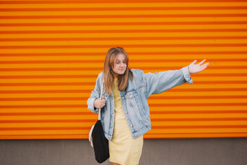 blonde teenager girl in a yellow dress and jute stands near an orange wall and spreads her arms.