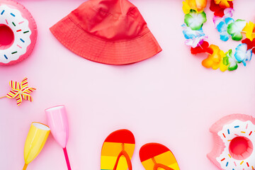 Flat lay of summer vibes concept with colorful pool party items, cocktail glasses, donut inflatable drink holders, flip flops, bucket hat and flower necklaces on pink background with copy space.