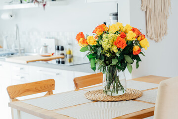 Home kitchen counter table with selective focus on on vase with yellow and orange roses on it with blurred background of modern cozy white kitchen. Home interior design details. Copy space.