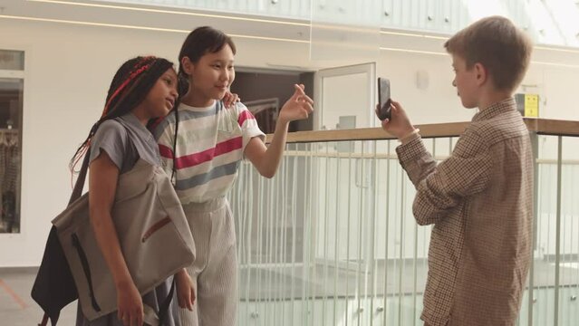 Two African American and Asian gen z girls posing for camera while their Caucasian classmate with smartphone in hand taking pictures of them at modern school corridor