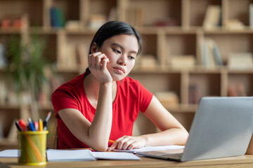 Boring sad adolescent asian female student watching video lesson, lecture on computer in room interior