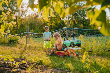 Cute little boys playing together on green summer garden background. Brothers riding plastic colorful cars. Family, happy childhood, friendship concept.