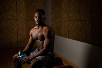 Fototapeta na wymiar Shirtless african american man drinking from a shaker in the gym.