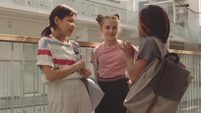 Medium shot of three diverse 12 year old school girls in casual clothes with backpacks chatting in modern school corridor with glass railings