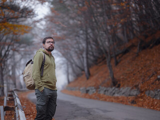 Young handsome man posing in autumn forest on road. young hipster guy with backpack , traveller standing in woods, Hiking, Forest, Journey, active healthy lifestyle, adventure, vacation concept.