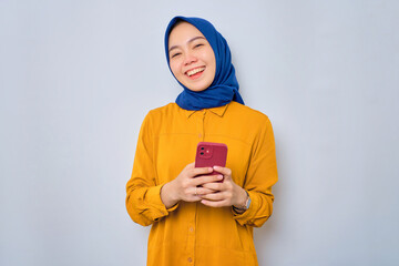 Smiling young Asian Muslim woman dressed in orange holding mobile phone and looking at camera isolated over white background