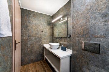 Modern bathroom in grey and brown colors with white bowl sink, tap mounted in the wall, square mirror. 