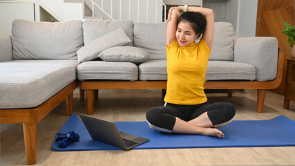 Fototapeta na wymiar Young woman exercising in bright living room and watching online tutorials on laptop
