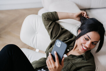 Cheerful attractive young African american woman sitting in cozy chair at home, having video call by phone in good mood. Successful entrepreneur working home with laptop, diaries on sofa. Happy girl.