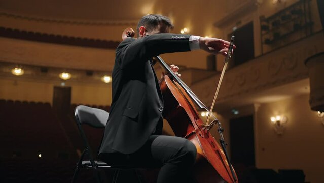 rehearsal of symphonic orchestra in philharmonic hall, cellist in black suit is playing cello on scene