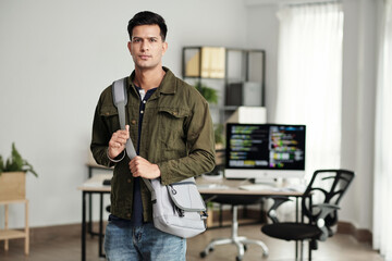 Serious young software developer with cross body bag standing against office background