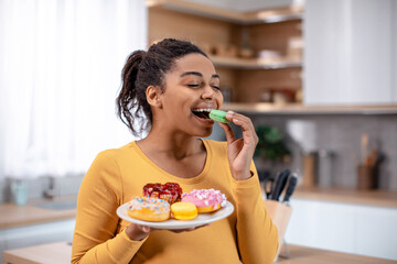 Glad millennial pregnant african american woman hold plate of sweets and eating cookies