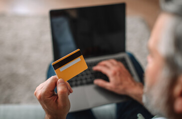 European senior male with beard looks at laptop with empty screen, shows credit card in living room