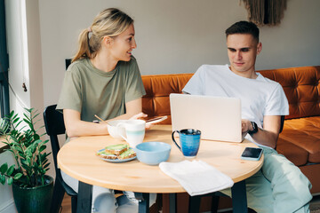 A man and a woman having breakfast use electronic gadgets and a laptop to surf the nets.