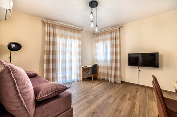 Simple modern living room interior with woodlike tile flooring, wooden desk at the corner, burgundy color sofa and TV set on  mounted on the wall. 