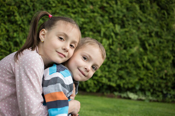 Portrait of charming smiling younger brother and older sister, sitting together in the park, hugging. happy lifestyle kids. Friendly relations between offspring