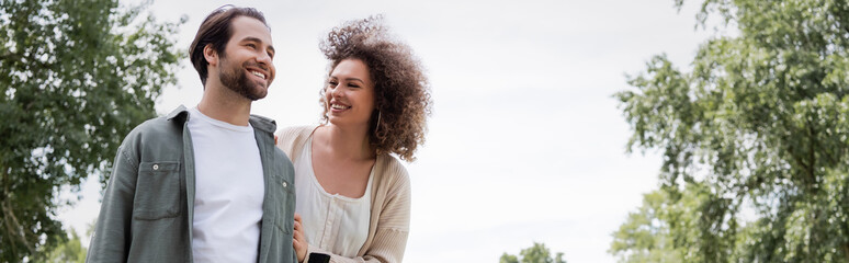 happy curly woman looking at man in summer clothes in green park, banner.