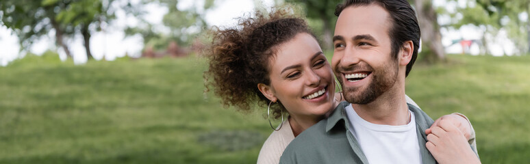 Fototapeta premium pleased and curly woman hugging boyfriend from behind in green summer park, banner.